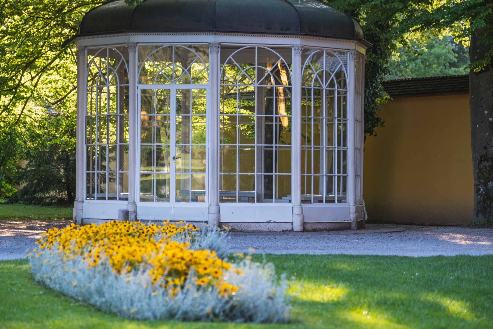 Ein weißer, verglaster Pavillon steht neben einer gelben Mauer, umgeben von grünem Gras und leuchtend gelben Blumen im Vordergrund.