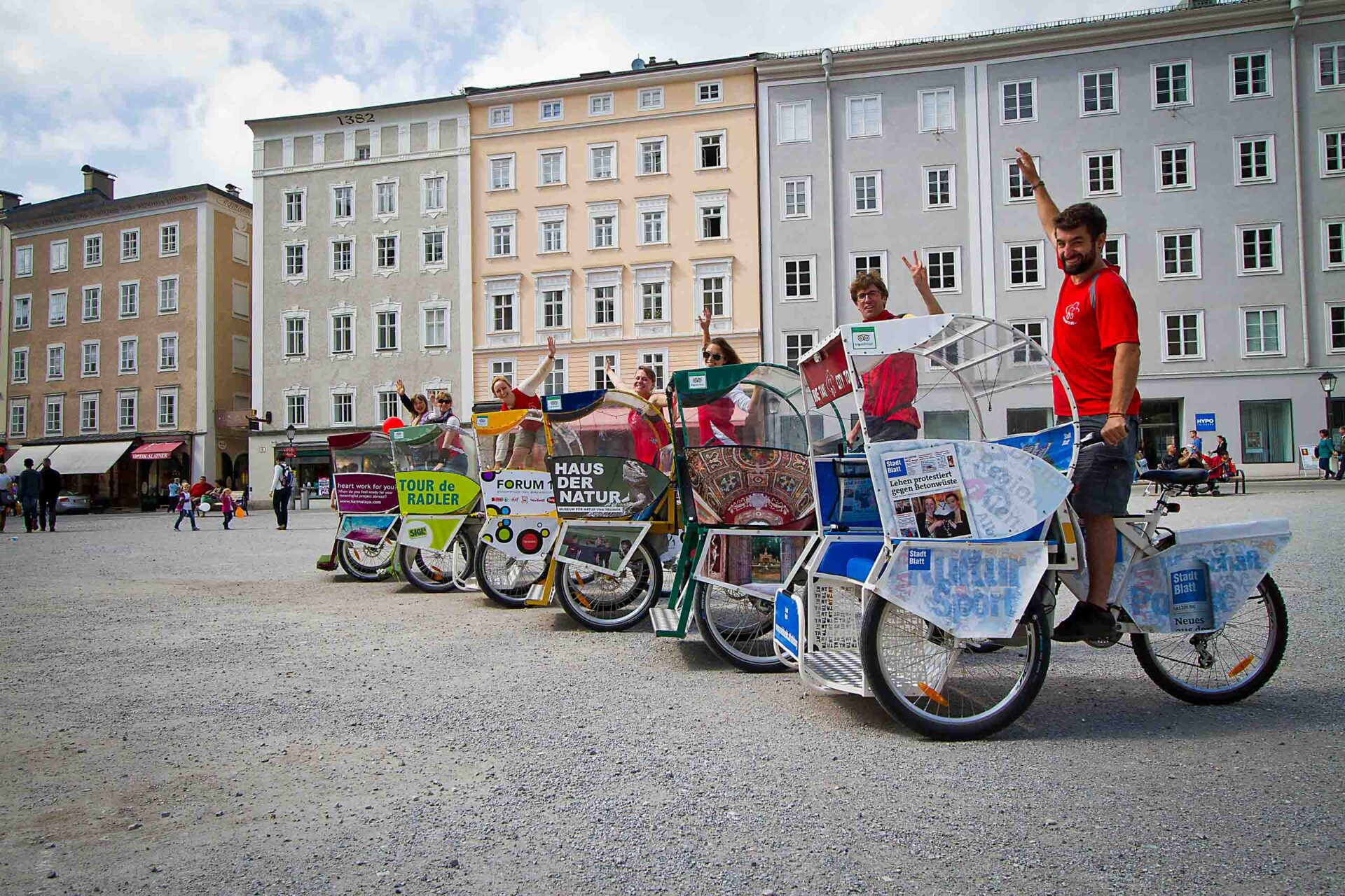 Eine Gruppe von Menschen posiert und winkt, während sie auf dekorierten Fahrrädern auf einem Stadtplatz mit historischen Gebäuden im Hintergrund sitzen.