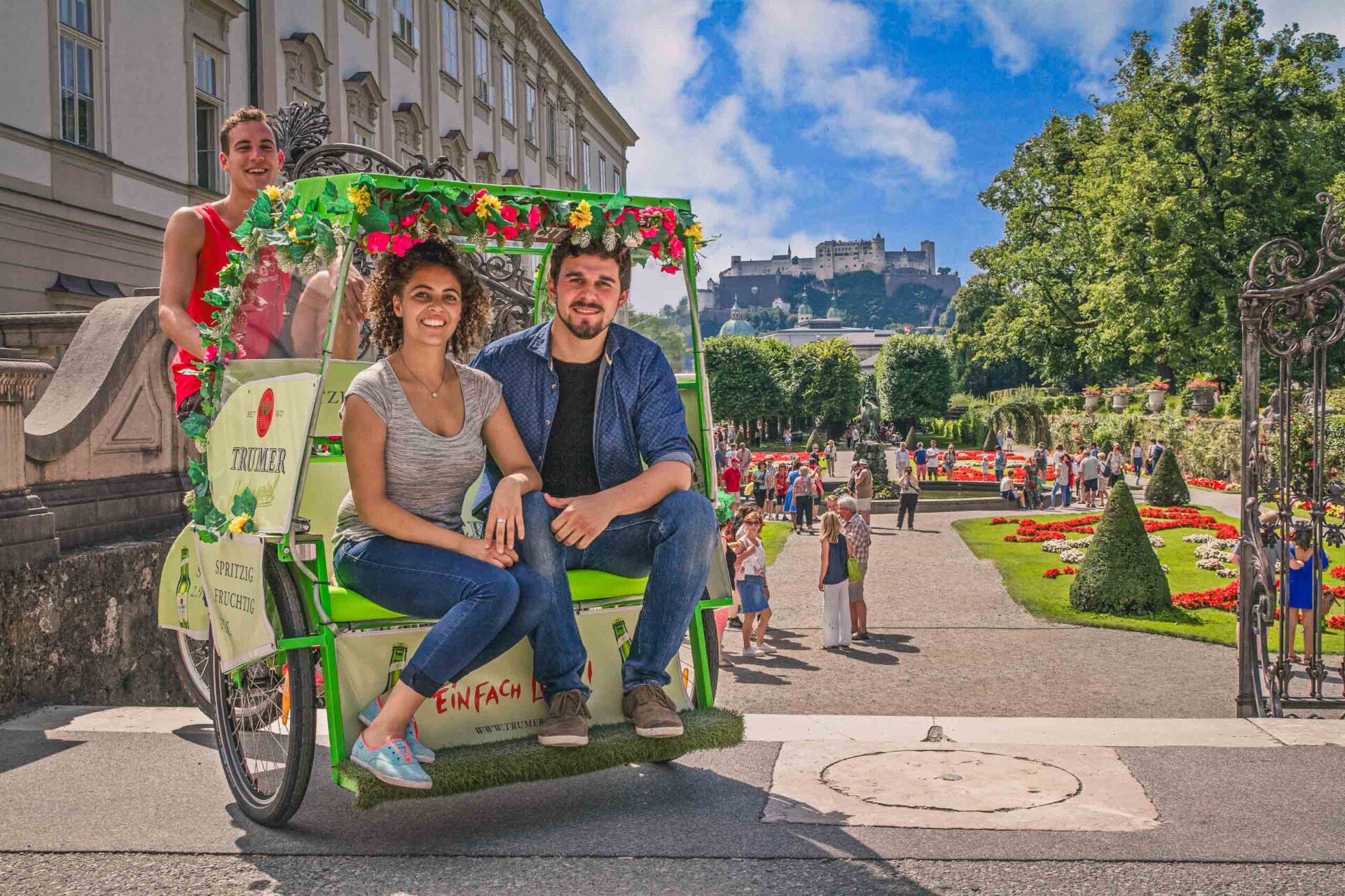 Drei Personen fahren in einer geschmückten Rikscha durch einen belebten Garten mit bunten Blumen, Wegen und einem großen historischen Gebäude und einer Festung im Hintergrund.