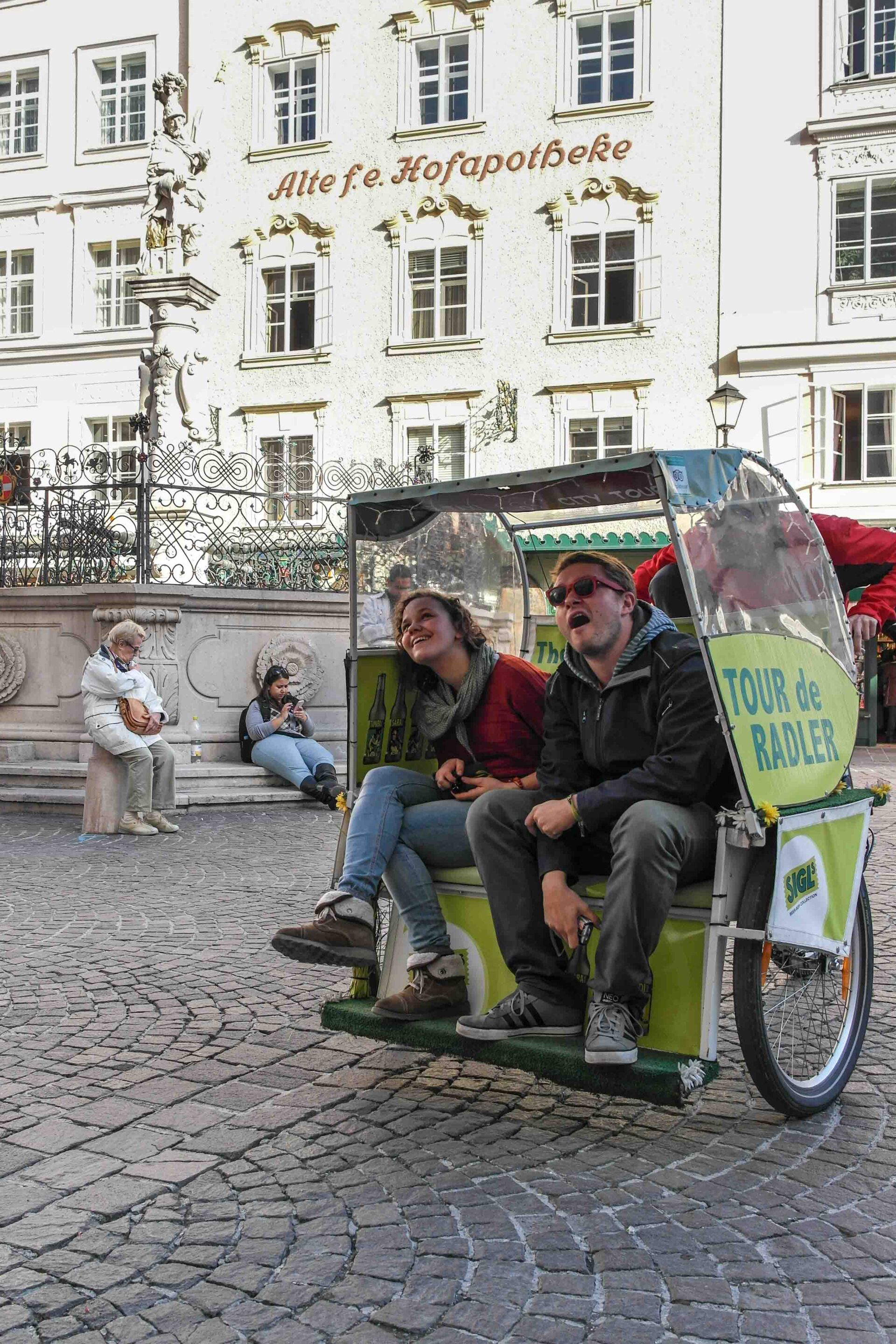 Zwei Personen sitzen in einem Fahrradtaxi mit der Aufschrift "Tour de Radler" auf einer Kopfsteinpflasterstraße, im Hintergrund sind andere Personen und historische Gebäude zu sehen.