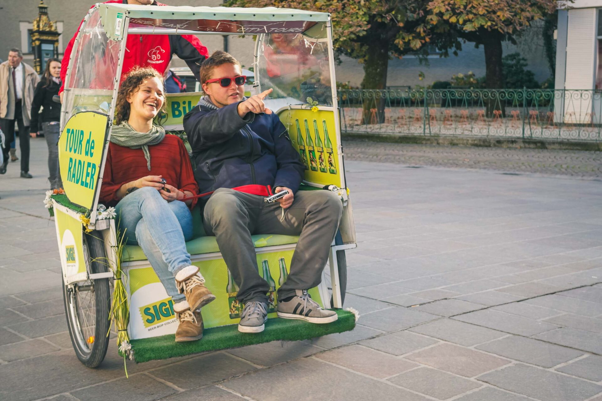 Zwei Personen sitzen in einem grün-gelben Fahrradtaxi auf einer gepflasterten Straße, wobei eine Person nach vorne zeigt und andere im Hintergrund gehen.
